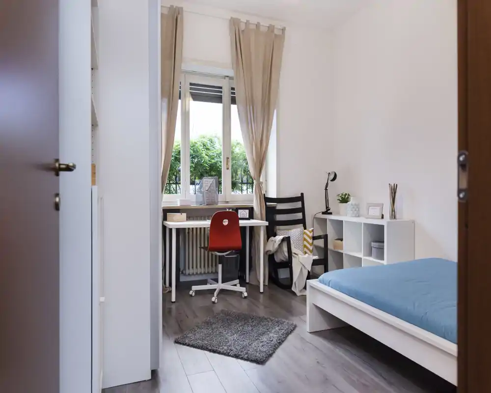 Bright, simply styled bedroom with a single bed, desk by the window and tasteful accessories. Natural light and soft tones create a calm workspace and sleeping area.