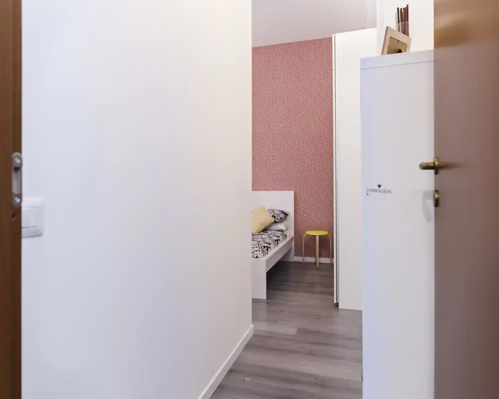 Pequeña habitación luminosa vista desde el pasillo: cama individual con ropa de cama estampada y cojín, papel pintado rosa decorativo y pequeño taburete.