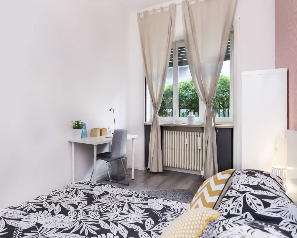 Dormitorio luminoso y bien decorado con colcha estampada, cojines decorativos y un pequeño escritorio junto a la ventana que deja entrar luz natural.