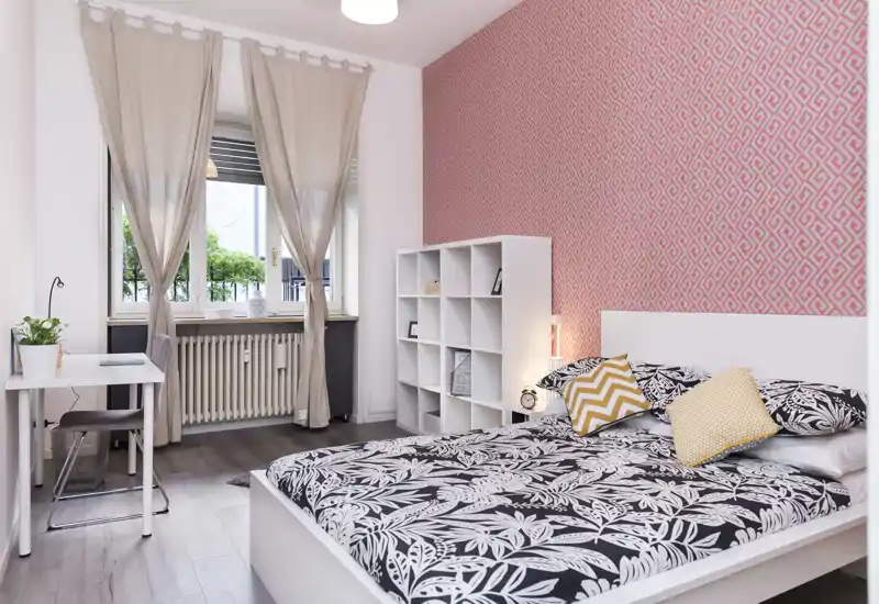 Dormitorio luminoso y moderno con pared decorativa, cama doble completa con cojines, estantería y pequeño escritorio junto a la ventana — ideal para estancias cómodas.