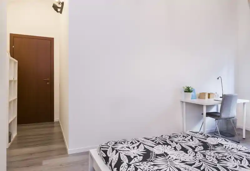 Dormitorio luminoso y minimalista con colcha de estampado, pequeño escritorio y silla junto a la pared, paredes neutras — acogedor y bien iluminado, ideal para estancias cortas.