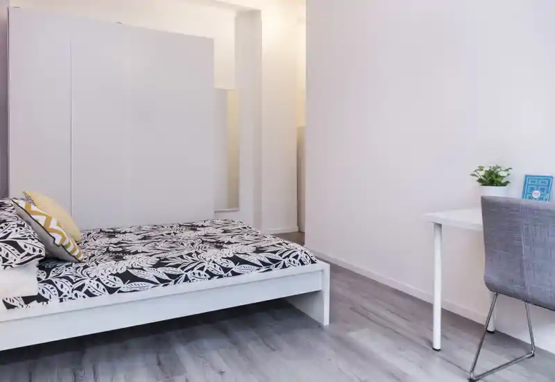 Dormitorio moderno y luminoso con una cama baja blanca, ropa de cama estampada y un pequeño escritorio. Líneas limpias y tonos neutros que crean un ambiente cálido, ideal para alquileres.