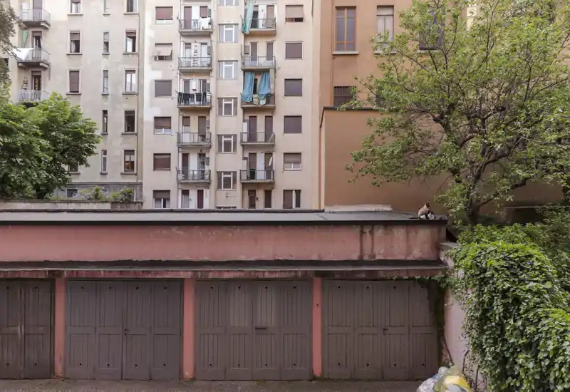 Vista esterna del cortile con una fila di box auto davanti a un edificio residenziale, con alberi e edera che aggiungono tocchi di verde.