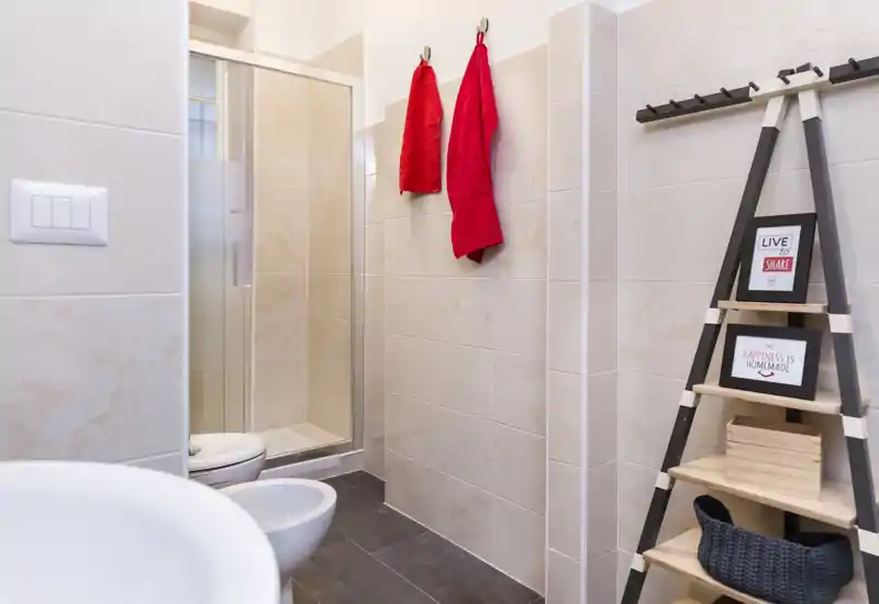 Bagno moderno e pulito con box doccia in vetro, bidet e WC, piastrelle neutre e asciugamani rossi che aggiungono un tocco vivace.