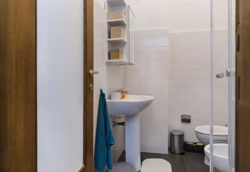 Bagno moderno e compatto con lavabo a colonna, WC, bidet e porta doccia in vetro. Mensole ordinate e un asciugamano turchese donano un tocco fresco.