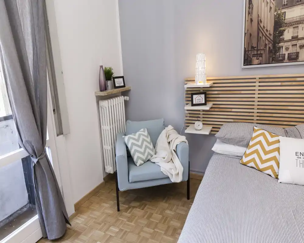 Angolo camera ben curato con letto comodo, cuscini decorativi e una poltrona vicino alla finestra che lascia entrare una luce naturale soffusa — perfetto per soggiorni accoglienti.