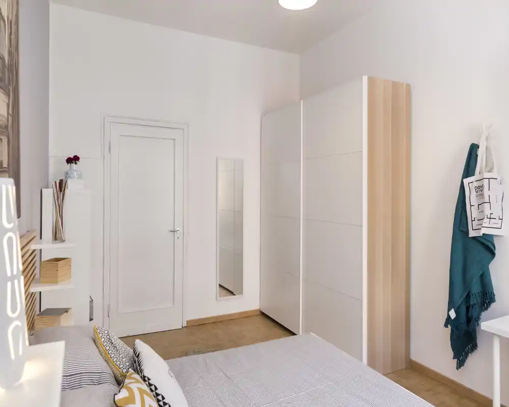 Camera da letto luminosa e moderna con letto ben rifatto, cuscini decorativi e un grande armadio bianco. Pavimento in legno caldo, specchio a figura intera e arredo essenziale e contemporaneo, perfetto per annunci di affitto.