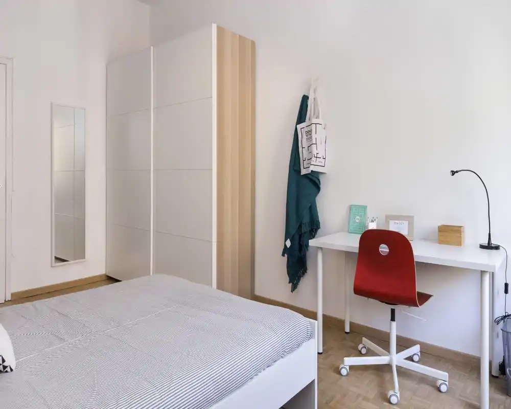 Camera da letto luminosa e minimale con un armadio bianco, letto ben rifatto in primo piano e una scrivania ordinata con sedia rossa—stile nordico pulito e buona luce naturale.