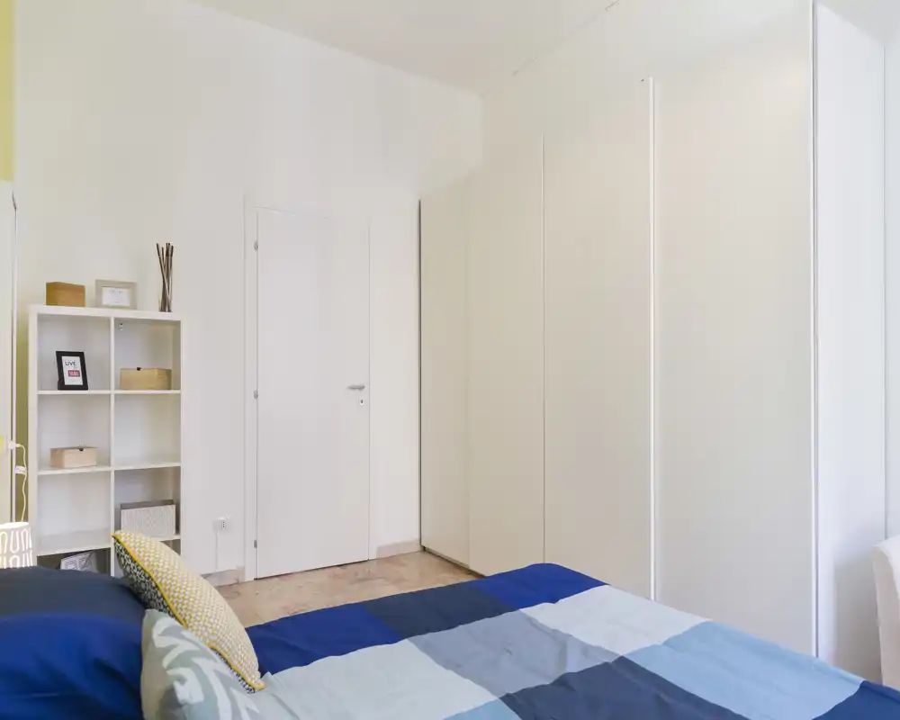 Dormitorio moderno y luminoso con cama doble bien hecha, amplio armario blanco y estantería minimalista — ideal para una estancia relajada.