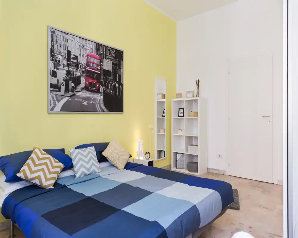 Dormitorio doble luminoso y bien decorado con una gran cama azul de cuadros, cojines decorativos y una fotografía de la ciudad encima de la cabecera. Una estantería blanca y lámpara de mesita crean un ambiente ordenado y acogedor, perfecto para anuncios.