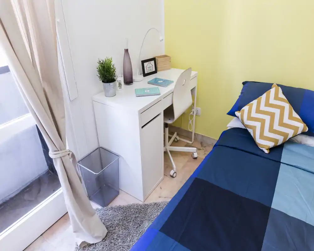 Rincón de dormitorio individual luminoso y ordenado con ropa de cama azul, cojín estampado y un escritorio blanco junto a la ventana — ideal para estudiante o viajero solo.
