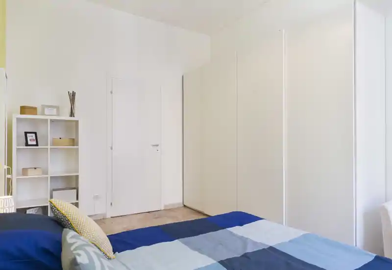 Camera da letto luminosa e ordinata con letto matrimoniale in primo piano, grande armadio bianco e libreria: stile minimal e pulito, ideale per un annuncio di affitto.