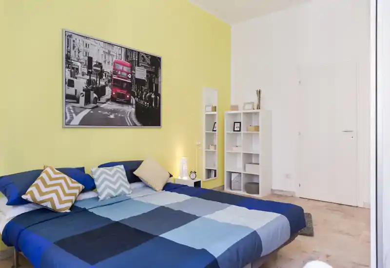 Dormitorio doble luminoso y bien decorado con una gran cama azul de cuadros, cojines decorativos y una fotografía de la ciudad encima de la cabecera. Una estantería blanca y lámpara de mesita crean un ambiente ordenado y acogedor, perfecto para anuncios.