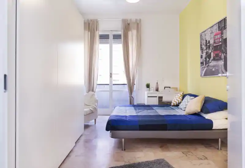 Dormitorio moderno y luminoso con cama doble con ropa de cama azul, cojines decorativos, pequeño escritorio y luz natural desde la puerta balcón — limpio, confortable y bien presentado para un anuncio.