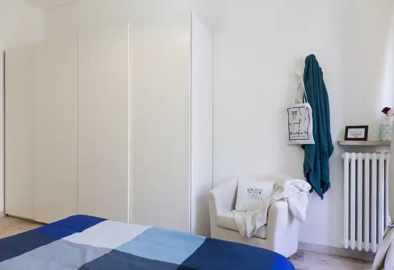 Dormitorio luminoso y moderno con cama bien hecha en tonos azules, un gran armario blanco y un sillón acogedor con mantas. Estilo minimalista y mucha luz natural lo hacen atractivo para huéspedes.