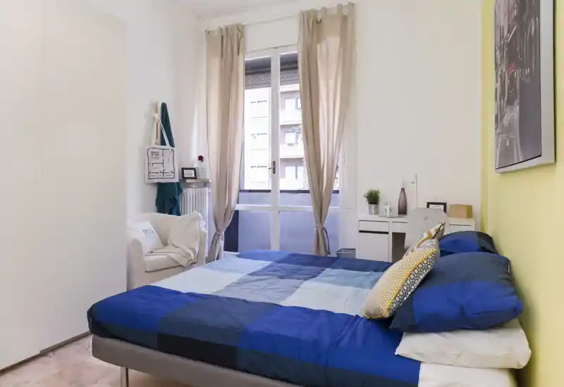 Dormitorio luminoso y bien decorado con cama doble con ropa de cama azul geométrica, un pequeño escritorio junto a la ventana y un sillón — ideal para una estancia cómoda con luz natural.