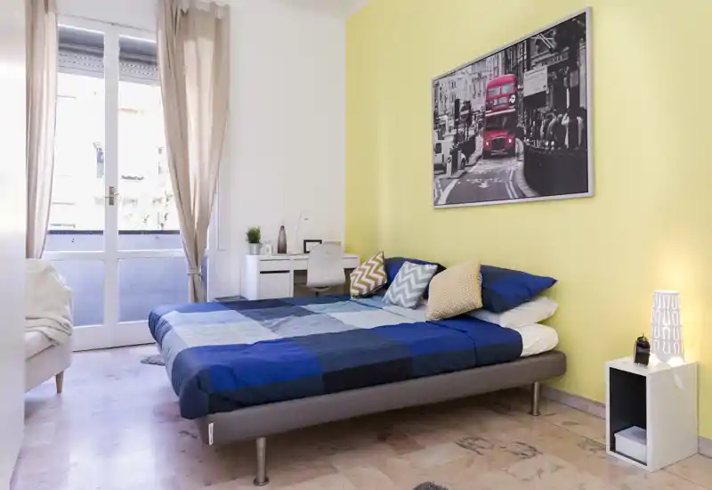 Dormitorio moderno y luminoso con cama doble vestida con ropa de cama a cuadros azules, cojines decorativos, lámpara de mesita y un pequeño escritorio junto a la puerta del balcón — ambiente fresco y acogedor.
