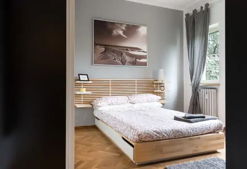 Dormitorio luminoso de estilo minimalista con cabecero de listones de madera, cama doble con ropa de cama estampada y ventana que aporta luz natural.