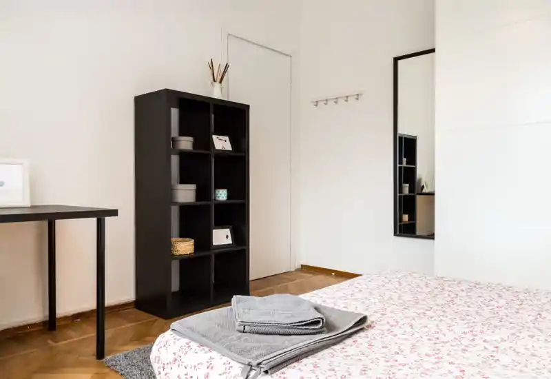 Dormitorio luminoso y minimalista con colcha estampada, toallas dobladas sobre la cama y una estantería negra. Paredes neutras y un espejo de cuerpo entero amplían la sensación de espacio.