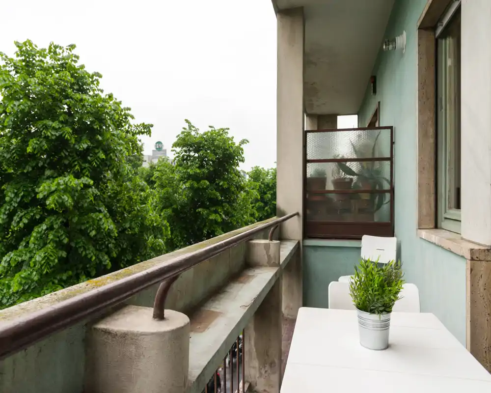 Sunny city balcony with a small table and potted plant, overlooking lush trees — a quiet spot for coffee or reading.