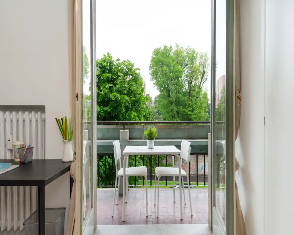 Bright balcony with a small white table and chairs, opening to leafy green views — ideal for morning coffee or a quiet outdoor meal.