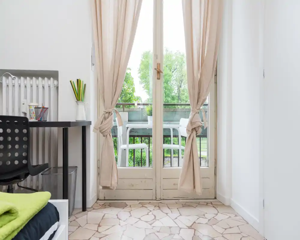Bright small bedroom corner with a glass door leading to a balcony. Neutral curtains, a bedside desk and a glimpse of the bed create a fresh, airy atmosphere ideal for a single guest.