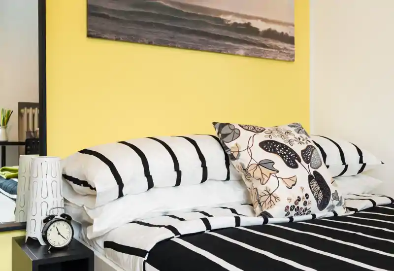 Bright, well-styled bedroom corner with a neatly made bed, patterned cushions and a modern side table with alarm clock. The yellow accent wall and striped bedding create a fresh, inviting look ideal for rental listings.
