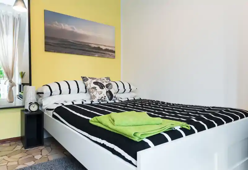 Bright, well-staged bedroom with a large double bed dressed in black-and-white striped linens, decorative pillows and neatly folded green towels on top. A window with light curtains and a coastal wall print complete the cozy, airy atmosphere.