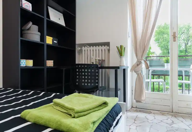 Bright, modern bedroom with a neatly made bed in the foreground, a black shelving unit and a small desk by a balcony door letting in abundant natural light.