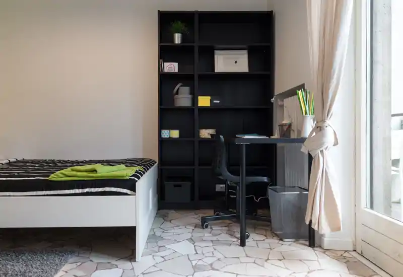 Bright, modern bedroom with a white single bed, striped bedding and a small study area next to a shelving unit and a window providing natural light.