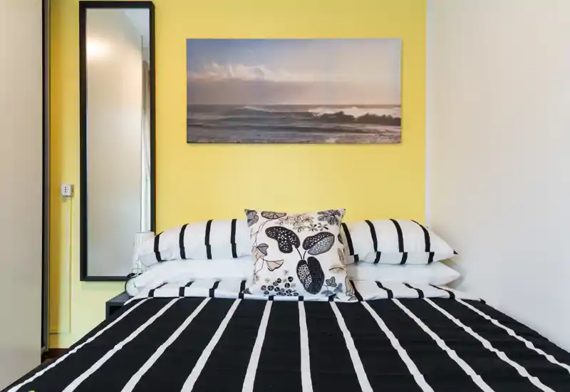 Bright, modern bedroom with a neatly made double bed, striped black-and-white bedding, decorative cushions and a seaside wall print above the headboard.