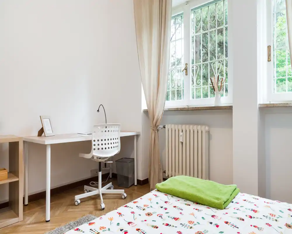 Bright, minimal bedroom with a patterned duvet, a neatly folded green towel, and a simple workspace by large windows letting in abundant natural light.
