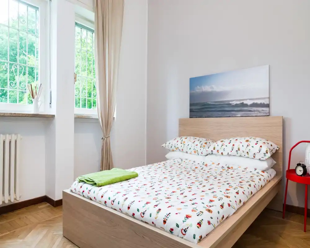 Bright, neatly staged bedroom with a wooden bedframe, patterned bedding and plenty of natural light from a large window.