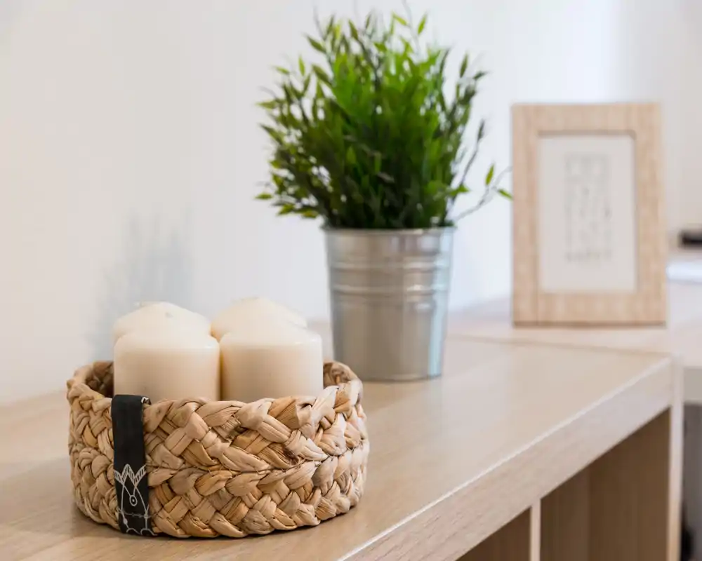 Close-up of a living area decorative vignette: woven basket with candles, a potted plant and framed print on a wooden shelf, creating a warm, minimalist accent.
