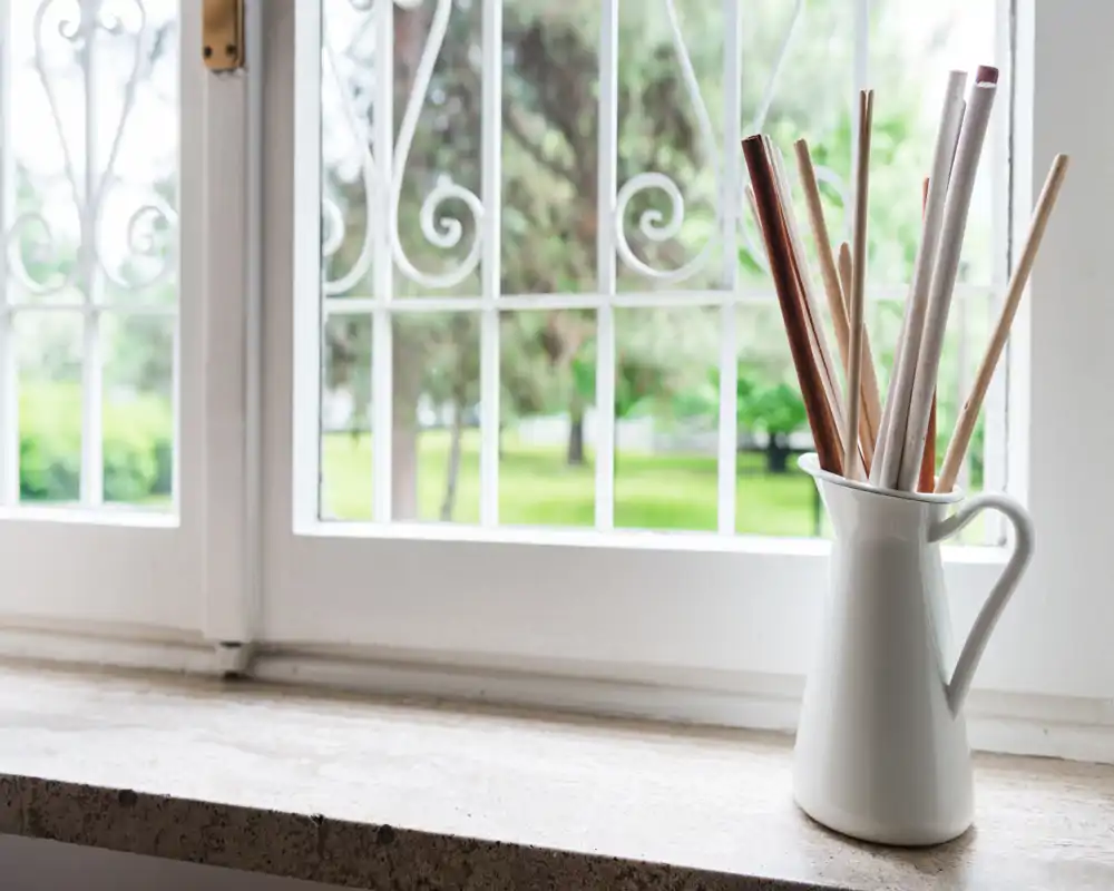 Bright window view overlooking a green outdoor area seen through decorative metal grilles; a simple white pitcher with reed sticks sits on the windowsill.