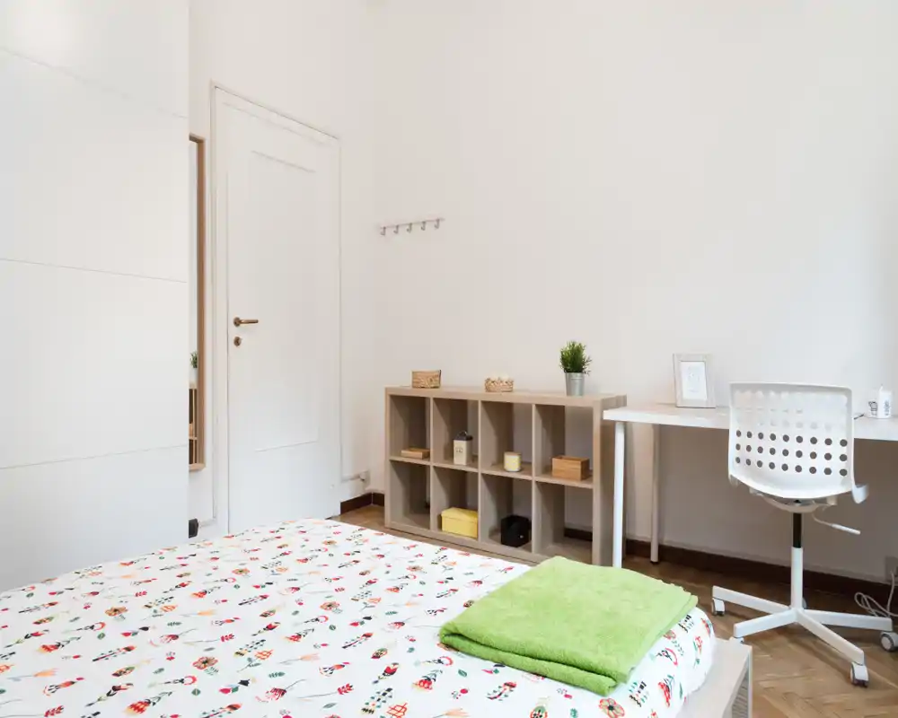 Bright, minimal bedroom with a patterned bedspread, simple shelving and a small desk — ideal for a single occupant seeking a tidy, functional space.