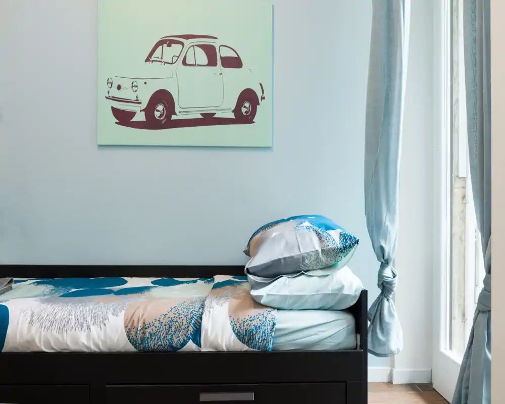 Bright, neatly styled single bedroom with patterned bedding, stacked pillows and a decorative car print above the bed; natural light from a window and soft blue tones create a calm atmosphere.