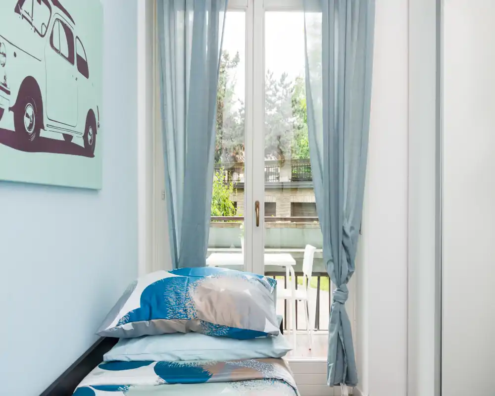 Bright single bedroom with a neatly made bed, soft blue textiles and a large glass door opening to a small balcony — ideal for a cozy, light-filled rental.