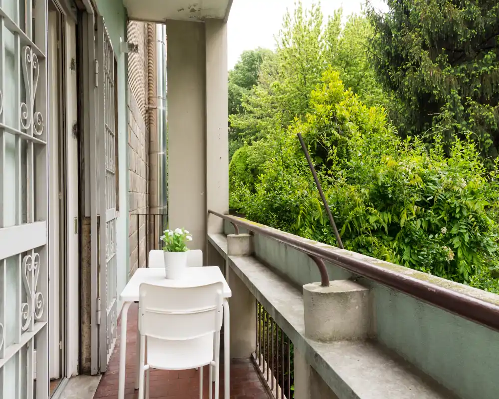 Sunny narrow balcony with a small white table and two chairs, overlooking lush green trees — ideal for morning coffee.