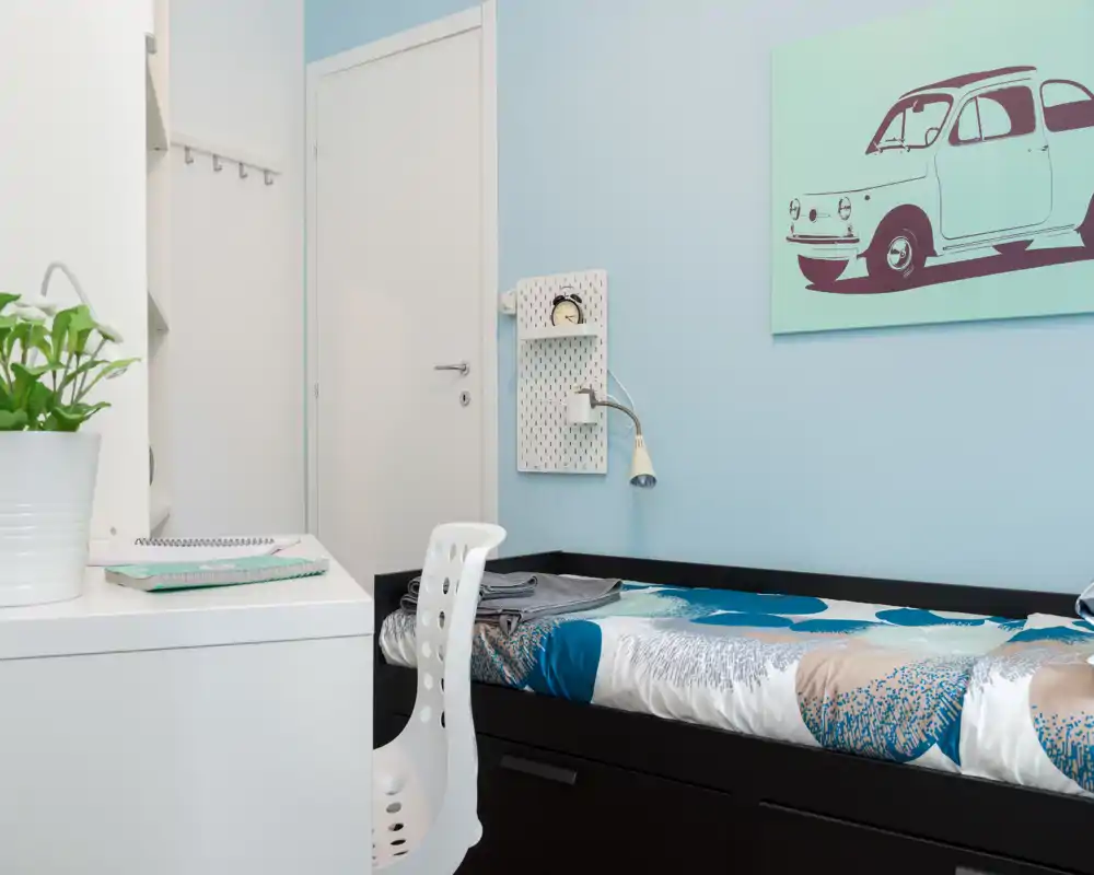 Compact, well-staged bedroom with a single bed, built-in storage drawers and a small desk; soft blue walls and coordinated bedding create a calm, welcoming atmosphere.