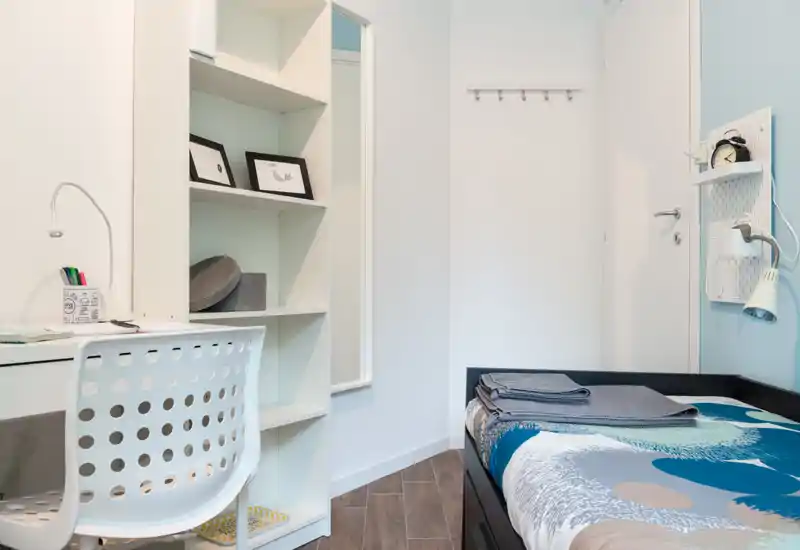 Compact, neatly styled bedroom with a single bed, bedside lamp and a small study desk. The room is bright, clean and efficiently furnished for a student or short-term stay.