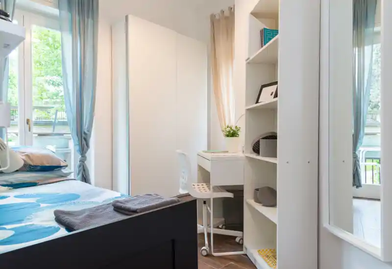 Sunny, well-styled bedroom with a double bed in the foreground, a desk and shelving by the window, and soft natural light creating a cozy atmosphere.