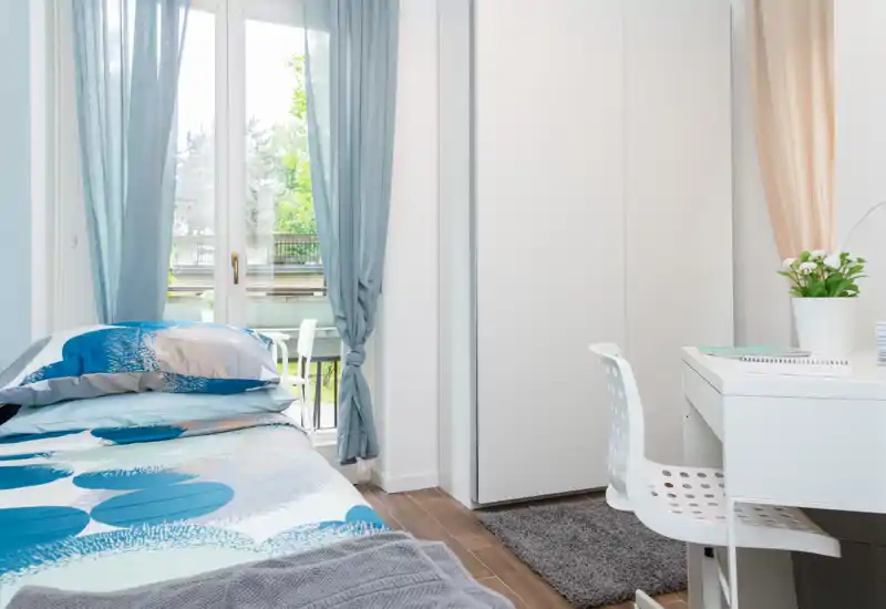 Bright, modern bedroom with a neatly made bed, desk area and large window letting in natural light; soft blue accents and simple wardrobe provide a calm, airy feel.