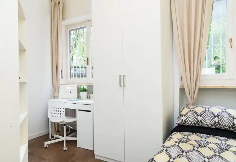 Bright, compact bedroom with a patterned single bed, white wardrobe and a small workspace by the window — ideal for a tidy student or short-term stay.