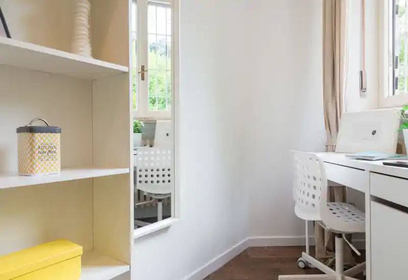Bright, compact home office corner with a white desk and perforated chair next to a window; clean shelving and soft curtains create a calm, airy atmosphere.