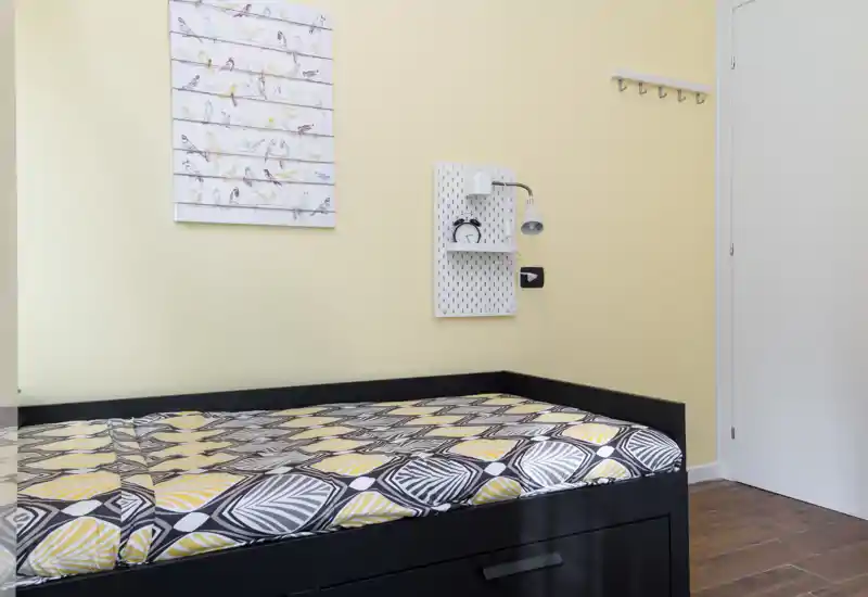 Compact single bedroom with a black daybed, patterned bedding and soft yellow walls; features a bedside wall lamp, shelf and framed bird artwork, ideal for a cozy one-person stay.