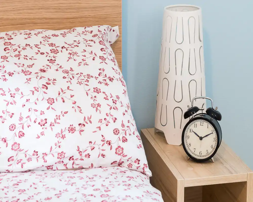 Gros plan lumineux d'une chambre montrant un oreiller à motif floral, une partie du matelas et de la tête de lit, une table de chevet avec lampe et réveil vintage — un coin nuit chaleureux et soigné.
