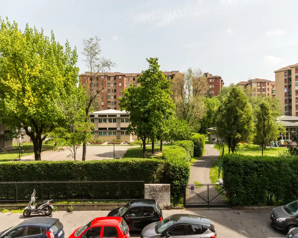 Espace extérieur commun avec arbres matures, haies et un chemin pavé traversant la zone verte; immeubles résidentiels visibles en arrière-plan.