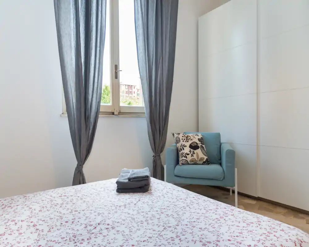 Chambre lumineuse et soignée avec un lit à motif floral au premier plan, un fauteuil bleu avec coussin décoratif et une grande armoire. La lumière naturelle de la fenêtre donne une ambiance calme et accueillante.