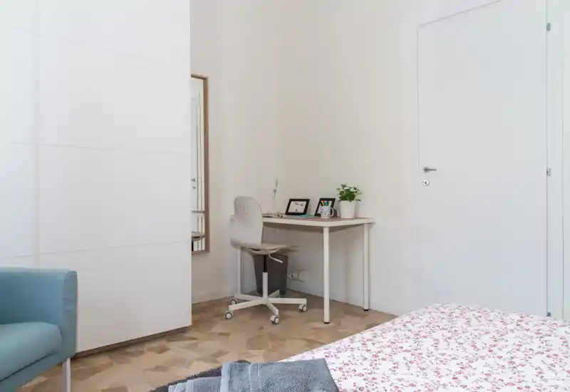 Coin chambre lumineux et minimaliste avec lit visible recouvert d'une couette à motifs, un petit bureau avec chaise et plantes, et une armoire. La photo met en valeur un espace nuit propre et bien éclairé, idéal pour une annonce locative.